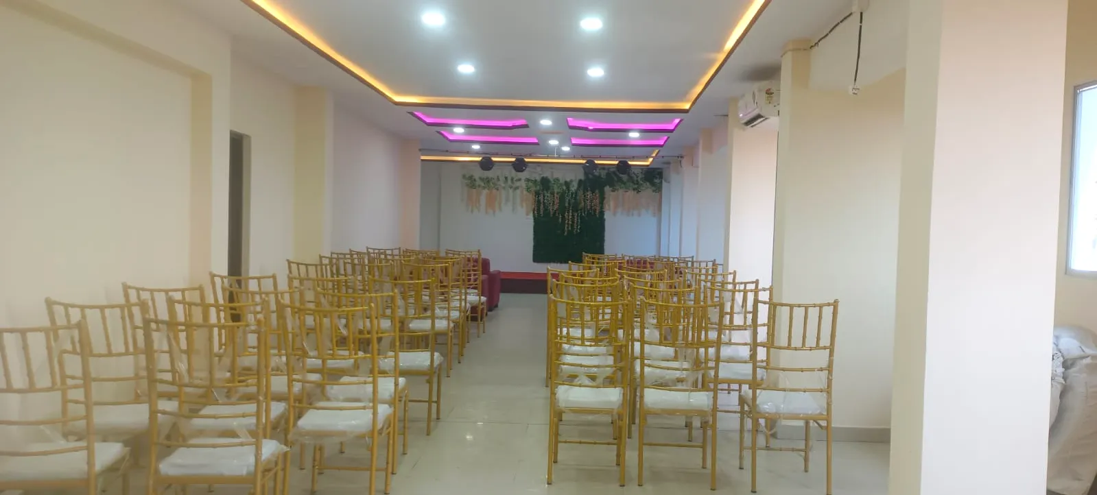 Banquet hall with rows of gold Chiavari chairs either side of an aisle leading to a greenery stage backdrop