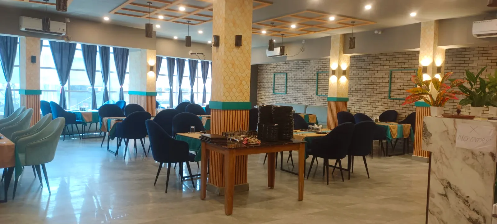 Neighborhood dining hall with navy and sea-green velvet chairs, navy curtains on tall windows, a brick wall and tiled pillars