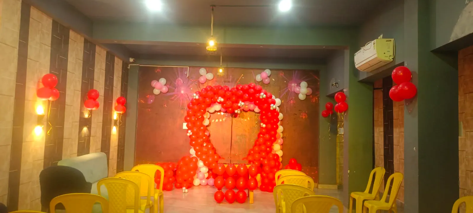 Party hall stage decorated with a red balloon arch, with yellow chairs in front